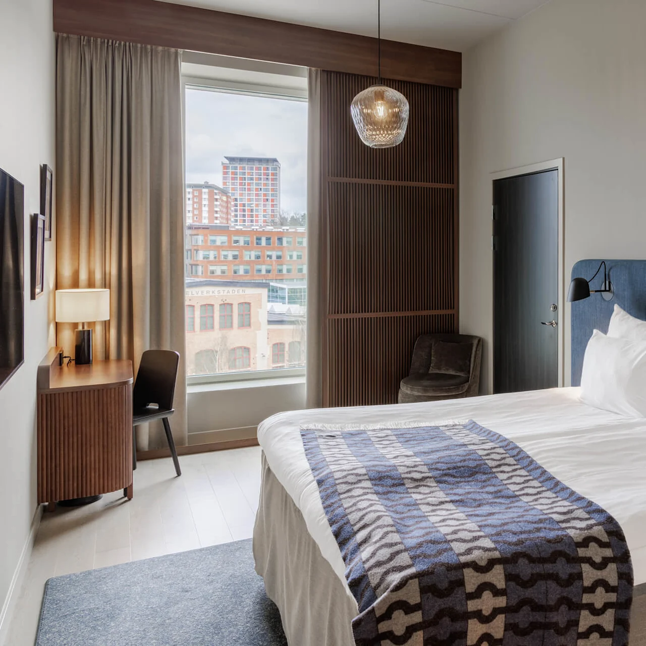A modern hotel room with a neatly made bed, city view window, and stylish furnishings. The decor suggests an upscale urban accommodation.