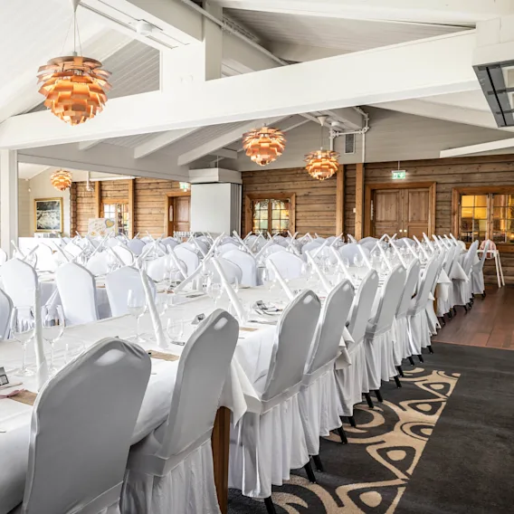 Long tables set with white linens and napkins fill a bright, spacious banquet room. Copper-colored pendant lights hang from the ceiling, and wooden walls add a rustic touch.