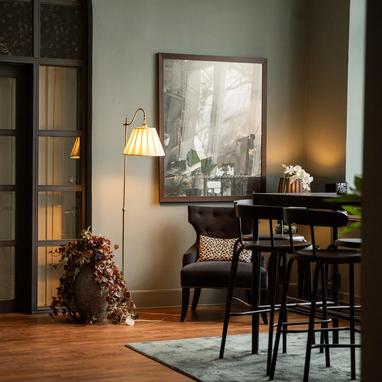 En golvlampa lyser upp ett mysigt rum med en svart fåtölj, dekorativ växt och högt bord med stolar. Ett stort inramat foto och glasdörrar skapar en inbjudande atmosfär.