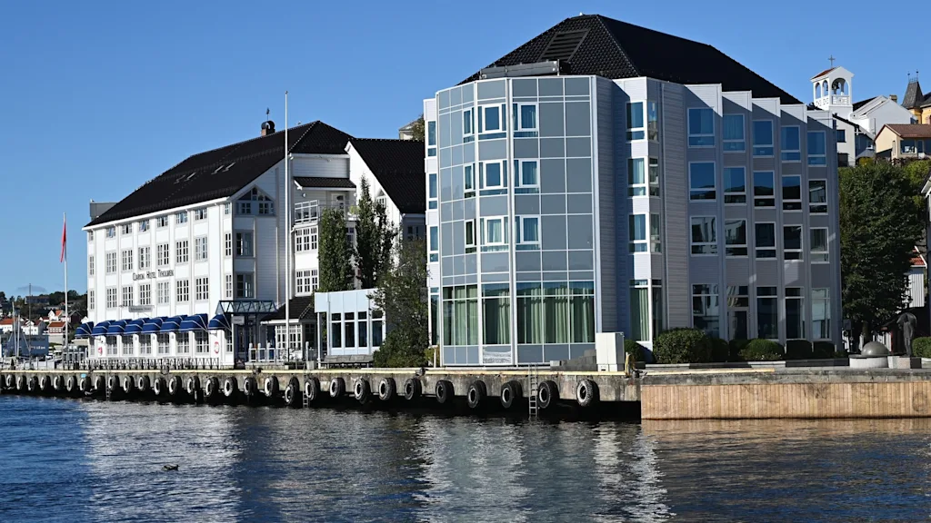 Et hotell med blå markiser og en moderne glassbygning ved vannkanten på en solrik dag. Teksten lyder Hotel.