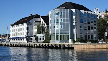 Et hotell med blå markiser og en moderne glassbygning ved vannkanten på en solrik dag. Teksten lyder Hotel.