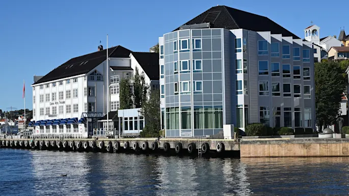 A hotel building with blue awnings and a modern glass building stand by a waterfront on a sunny day. Text reads Hotel.