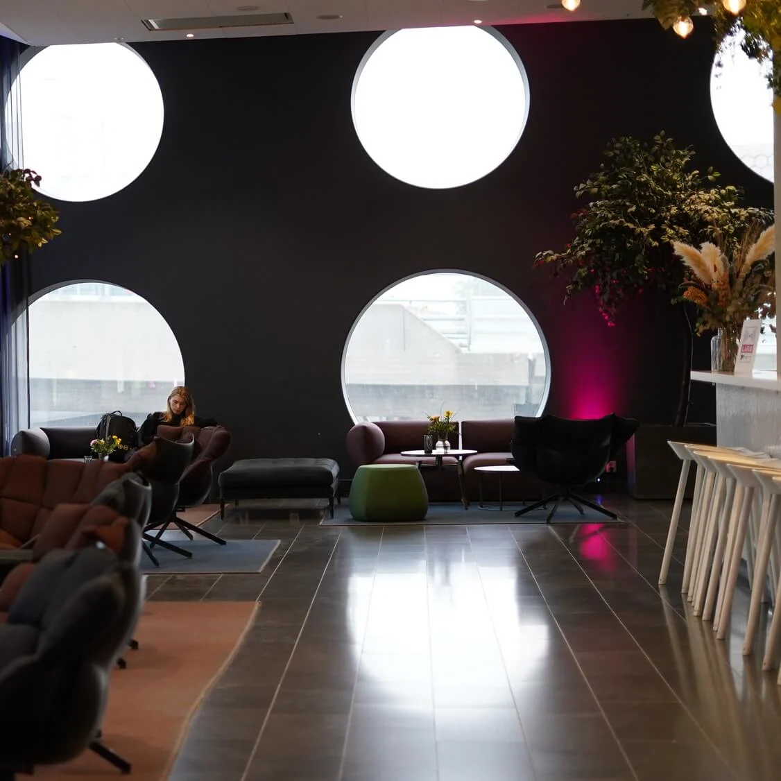 Komfortable siddepladser i en lounge; en person læser på en sofa. Store cirkulære vinduer og indendørs træer skaber stemning, mens bordene har vaser med blomster.