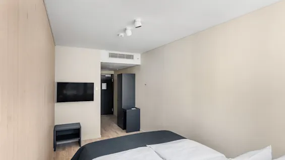Et moderne hotelværelse med en komfortabel seng, et vægmonteret fjernsyn og minimalistiske møbler, der skaber et rent og indbydende rum.