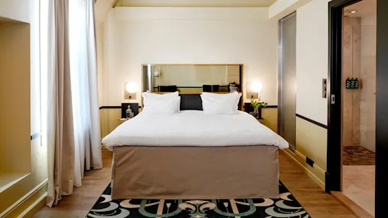 A neatly made bed with white linens is centered in a modern hotel room, flanked by side tables with spherical lamps. Large window with curtains to the left.