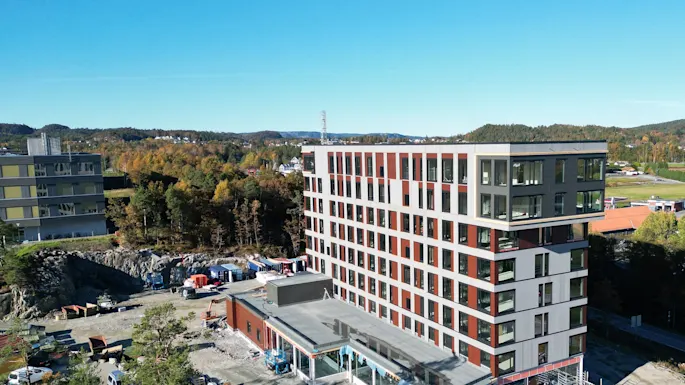 En moderne bygning under opførelse med klarblå himmel og efterårstræer i baggrunden, omgivet af byggeaktivitet.