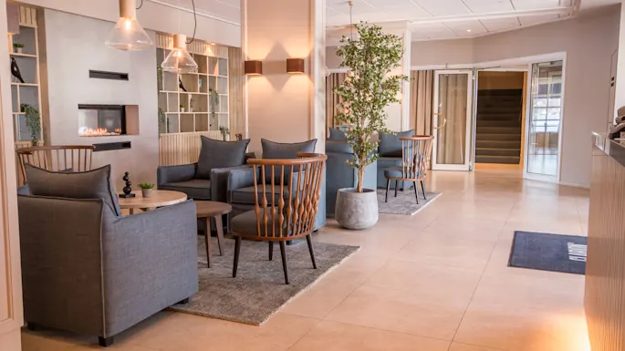 Elegant lounge area features grey armchairs and wooden chairs around round tables on rugs, with a fireplace in the background. Potted plants and soft lighting enhance the cozy atmosphere.