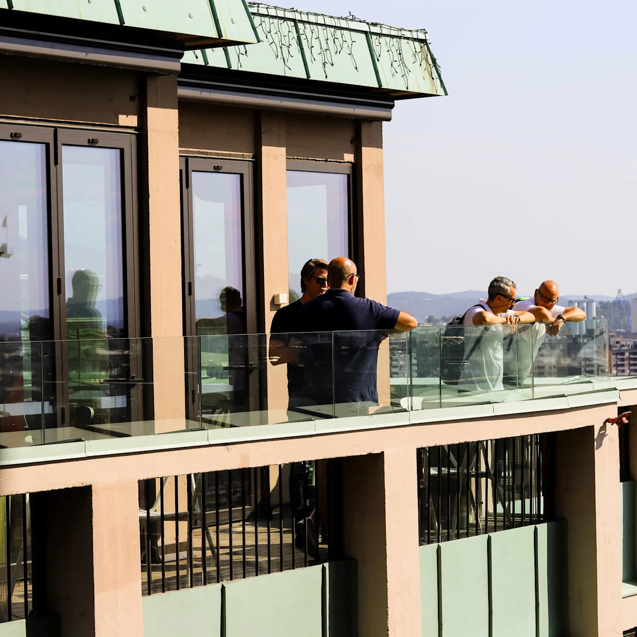 A group of people relaxes on a glass-paneled balcony of a modern building, overlooking a cityscape with a river and distant hills under a clear sky.