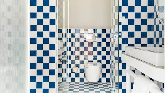 Et badeværelse har et hvidt toilet mod en væg med blå- og hvidternede fliser. En hvid vask er synlig til højre, suppleret af en glasbrusepartition.