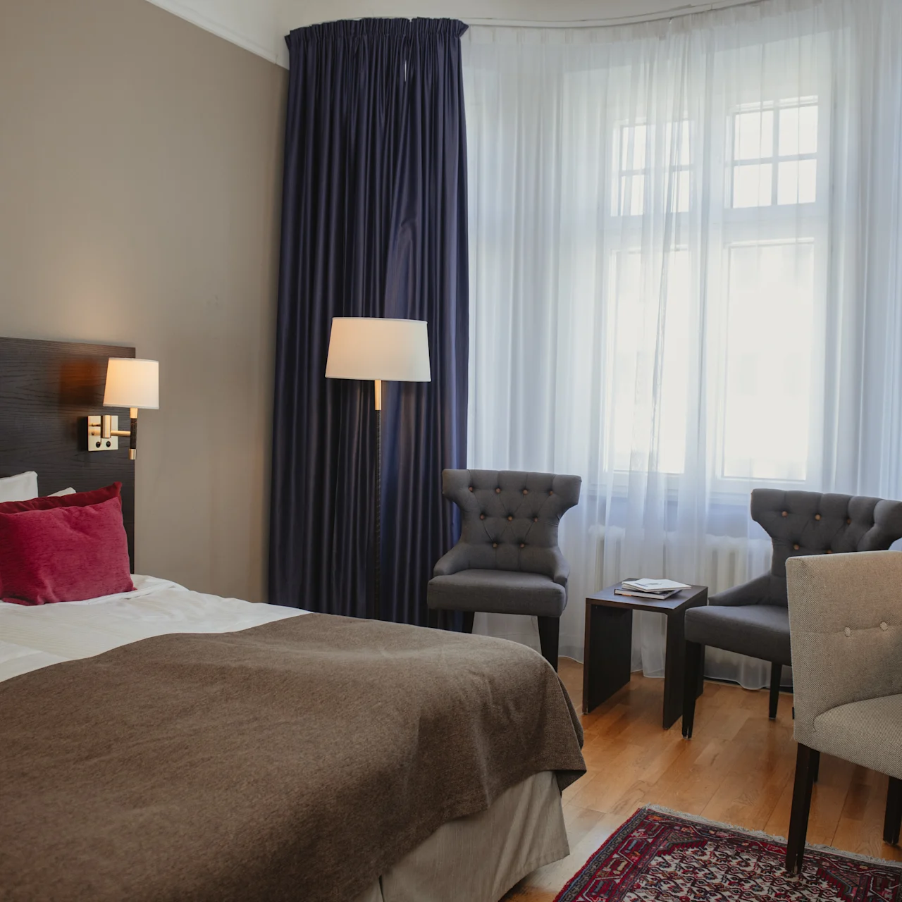 Dobbeltseng med brun dyne og røde puder i hotelværelse. To grå stole vender mod et lille bord ved et vindue med blå gardiner. Et skrivebord har en lampe og dekoration.