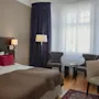 Dubbelsäng med brun bädd och röda kuddar i hotellrum. Två grå stolar vetter mot ett litet bord vid ett fönster med blå gardiner. Ett skrivbord har en lampa och dekorationer. 