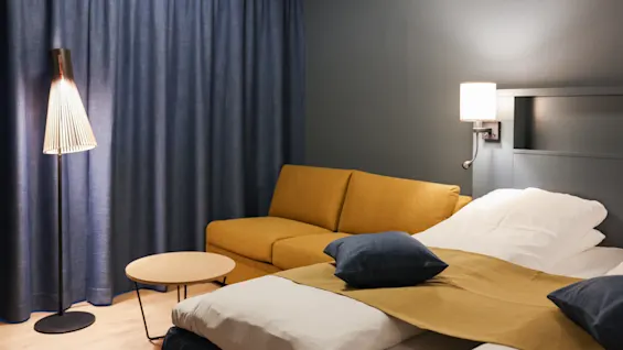 Säng med kuddar och filt, intill en gul soffa och runt bord i ett mjukt upplyst hotellrum. Blå gardiner täcker fönstret, med en golvlampa i närheten.