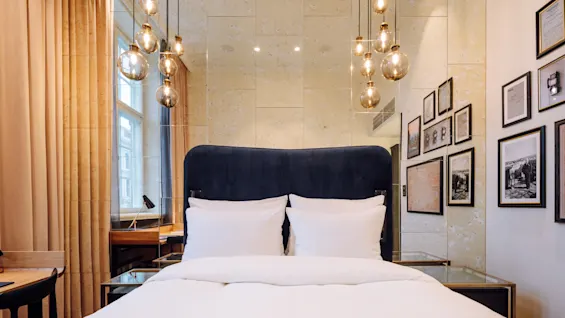 A luxurious bed with a dark headboard is set against a mirrored wall, adorned with hanging spherical lights. Framed pictures decorate the side wall. Soft curtains cover a window.