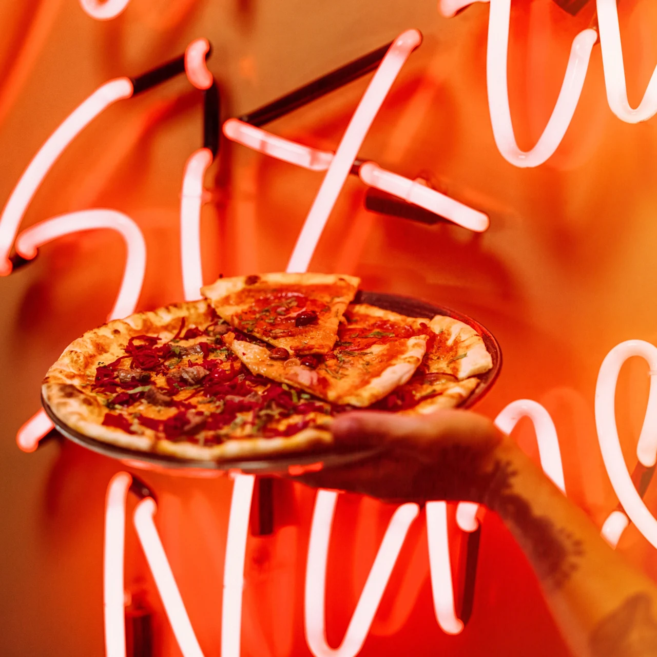 A hand holds a pizza with various toppings on a plate, set against a backdrop of bright, red-orange neon lights with cursive letters.