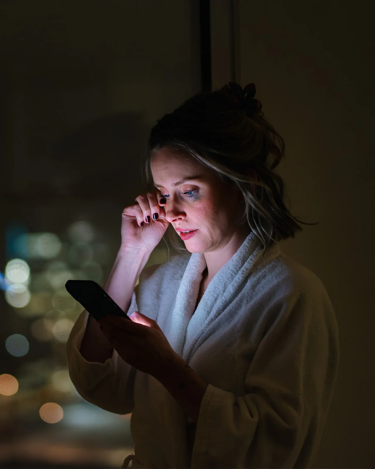 En person i badekåbe kigger på sin telefon om natten, byens lys sløret i baggrunden, et øjebliks stille eftertanke.