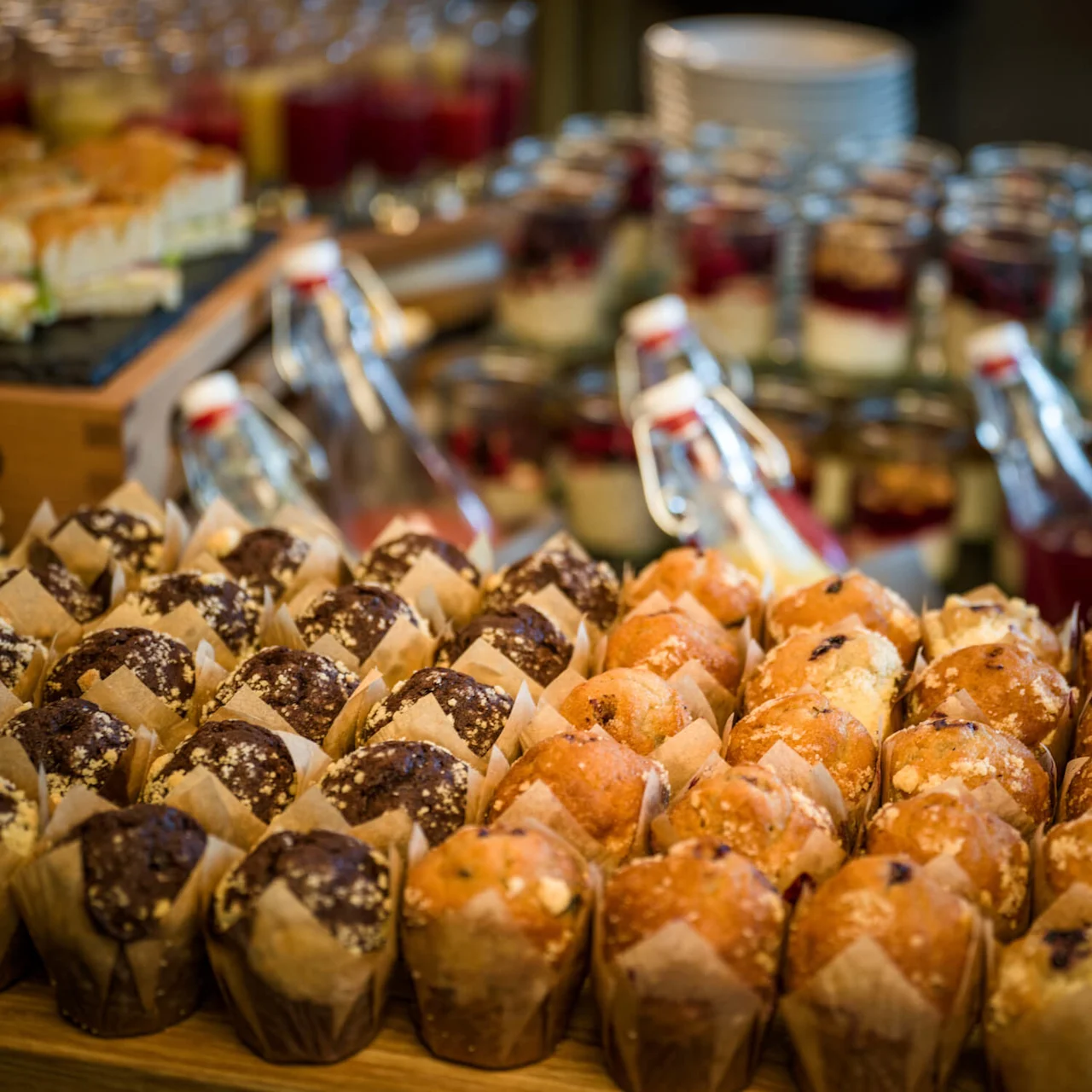 Muffins er pent plassert på et brett i tre; i bakgrunnen viser glassflasker og krukker med desserter og drikker en buffet.