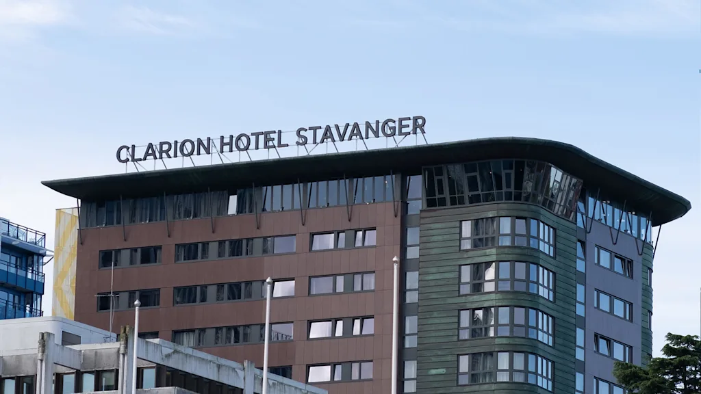 Clarion Hotel Stavanger-skylten sitter ovanpå en modern, flervåningsbyggnad med bruna och gröna paneler, omgiven av en klarblå himmel.