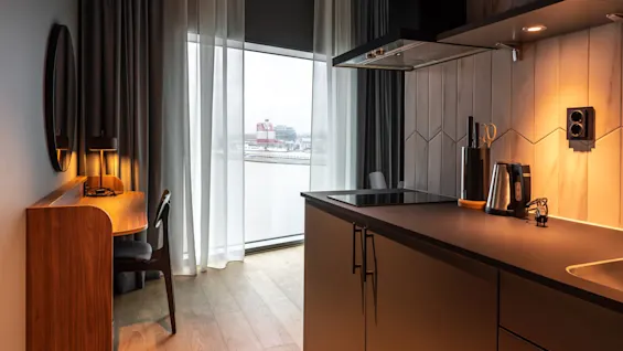 Ett mysigt kök med moderna apparater, inklusive en vattenkokare och spis, ligger intill ett fönster med utsikt över en vattendrag och stad. Ett skrivbord med lampa finns nära.