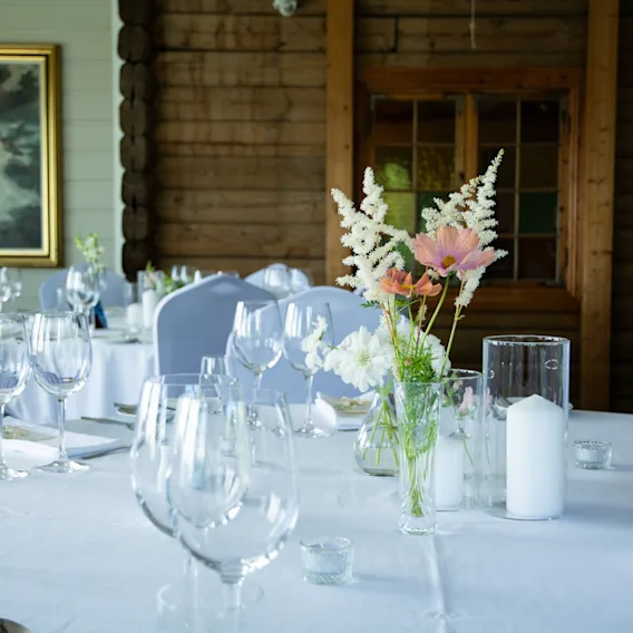 Nærbilde av borddekking til bryllup med klare vinglass, hvite kubbelys og en vakker blomsterdekorasjon.