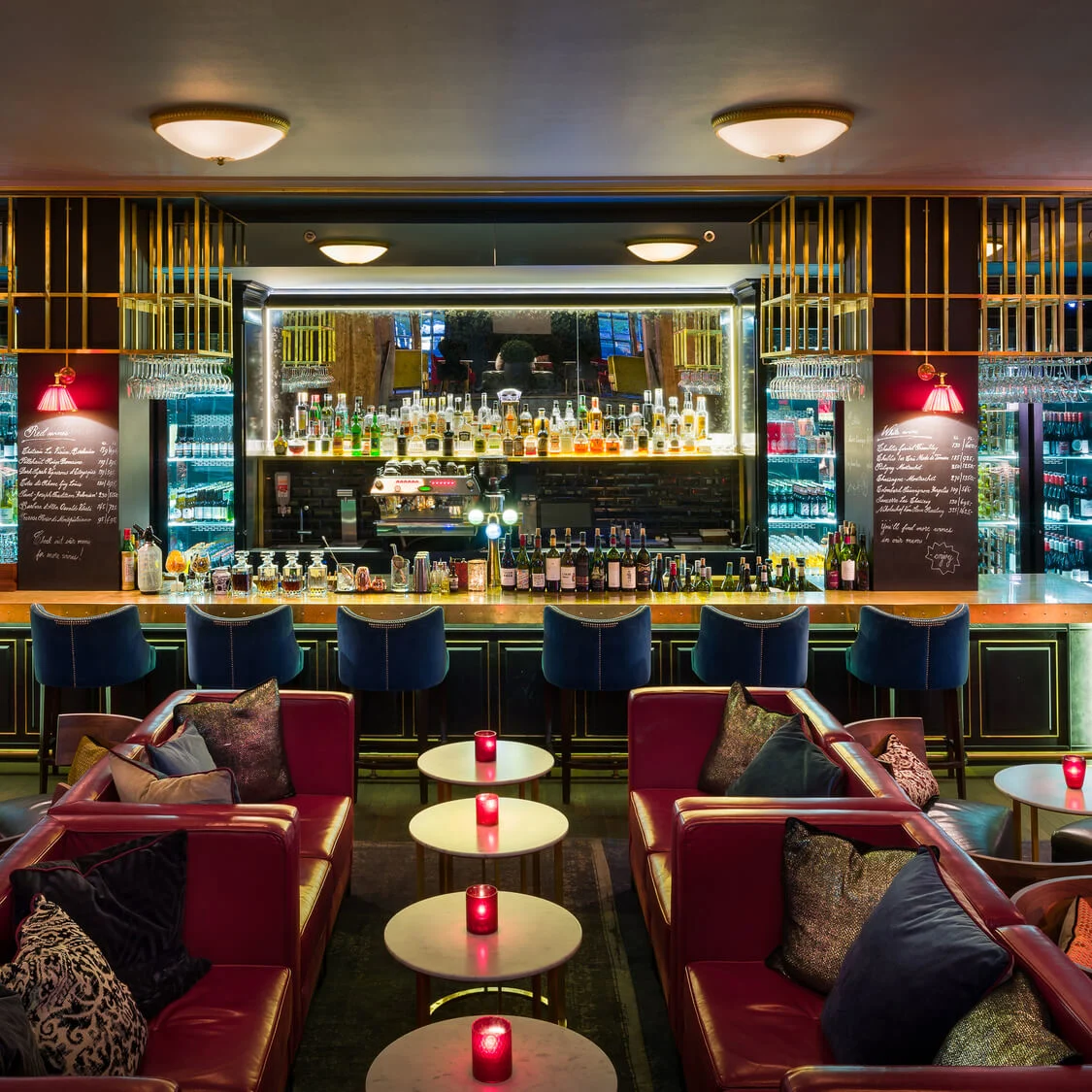 A stylish bar with bottles displayed, blue chairs, and red lanterns, surrounded by dim lighting and plush seating. Decorative skeleton lamps flank the interior.