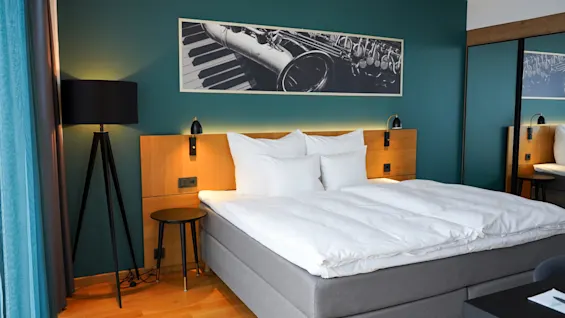 En pent seng står i et stilig hotellrom med grønne vegger, flankert av vegglamper og en gulvlampe. Over, et bilde med en saksofon og pianotangenter.