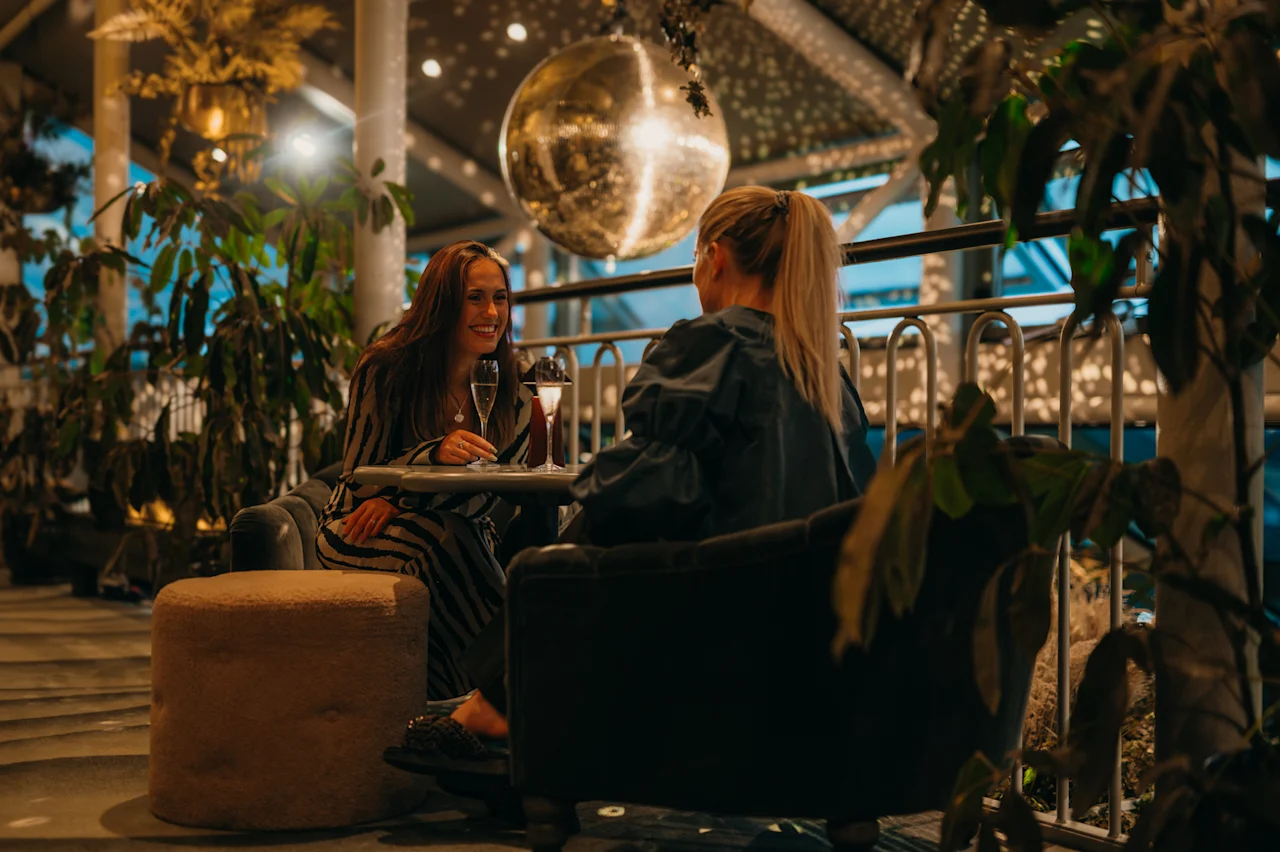 Kaksi ihmistä nauttii juomia ja keskustelua viihtyisässä, hämärästi valaistussa loungessa kasvien ja diskopallon alla.