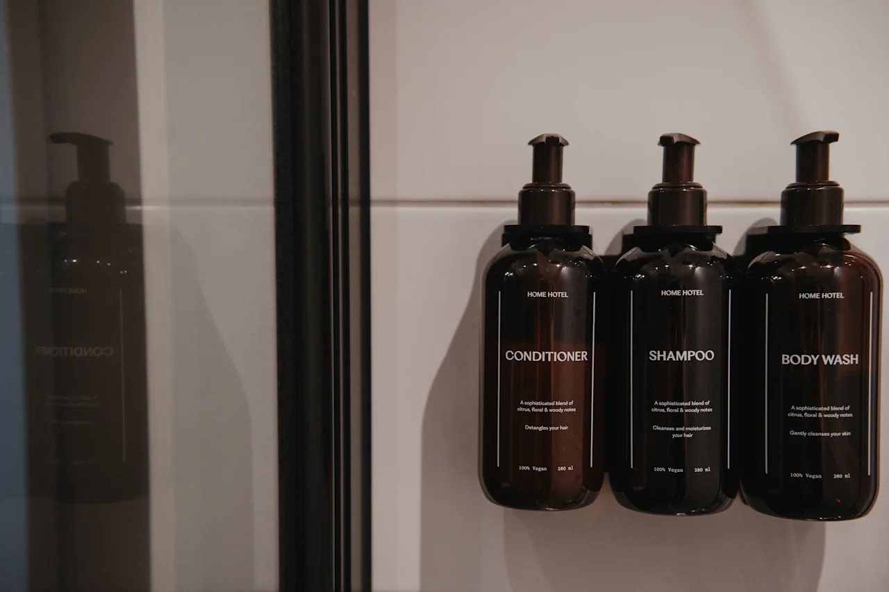 Tre mörka flaskor märkta Balsam, Schampo och Kroppstvål är monterade på en vit kaklad vägg i en dusch.