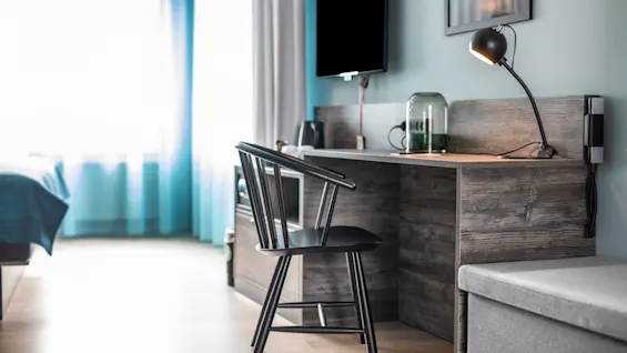 A wooden desk holds a lamp and glass jar while a black chair faces it. A TV is mounted on the wall. The room is softly lit with natural light.