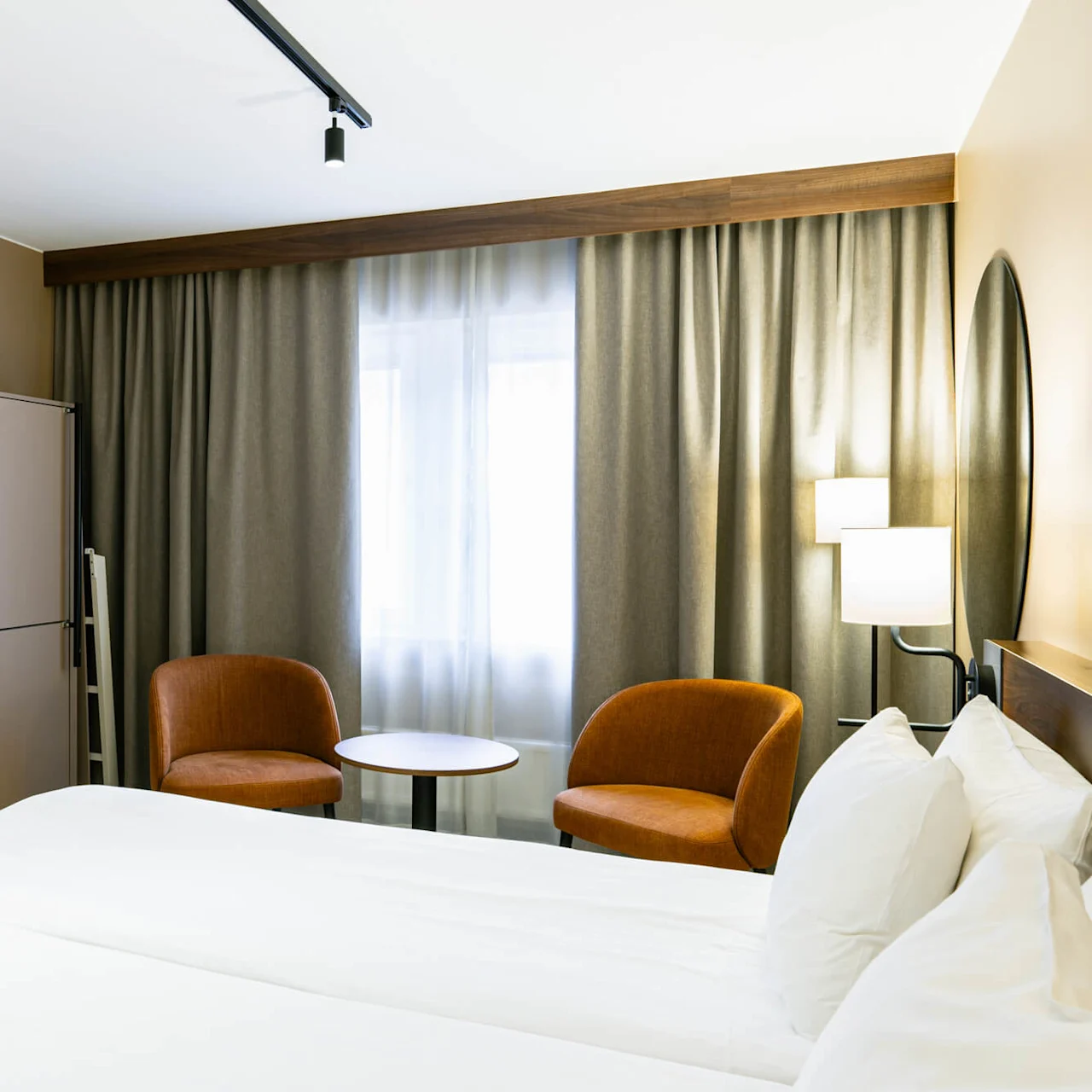 Seng med hvide lagner; to brune stole og et rundt bord ved vindue med grå gardiner; væglampe ved siden af et ovalt spejl skaber en hyggelig hotelstemning.