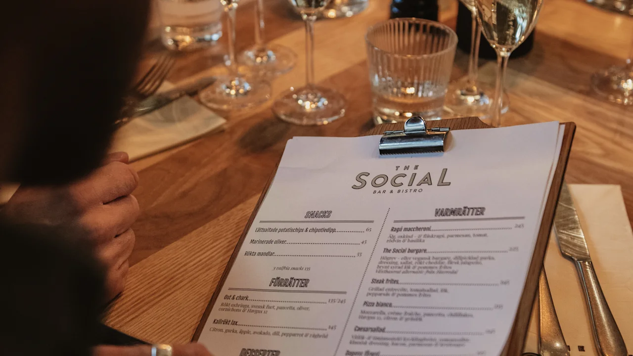 A person holding a menu labeled "The Social Bar & Bistro," seated at a table set with wine glasses, utensils, and a lit candle in a restaurant environment.