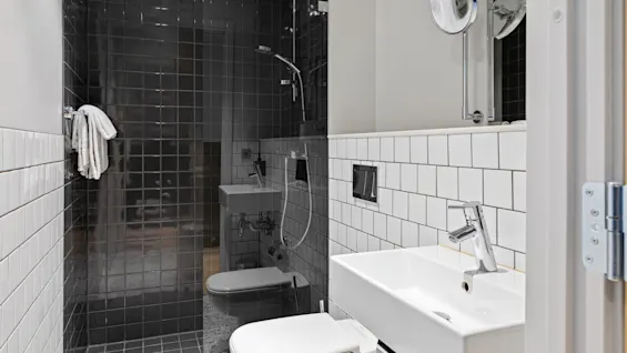 Et moderne bad med hvit vask og toalett; et stort speil dekker veggen over. Dusjområde med mørke fliser inkluderer en montert dusjhode. Hvite fliser på midtveggen.
