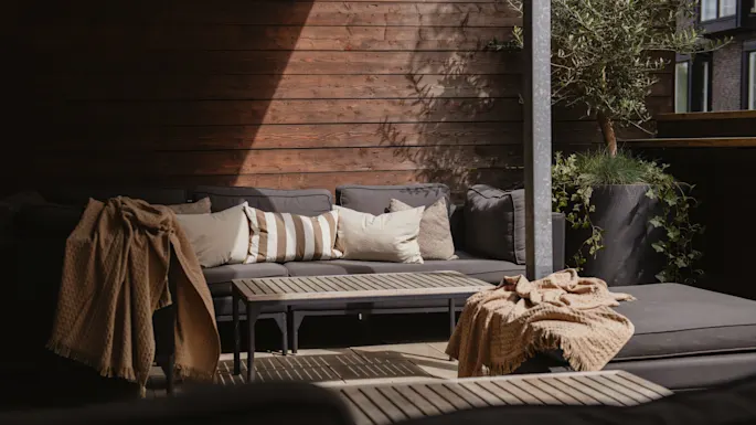 Et hyggeligt udendørs siddeområde med grå sofaer med beige puder, træborde og dekorative tæpper, omgivet af et træhegn og store potteplanter.