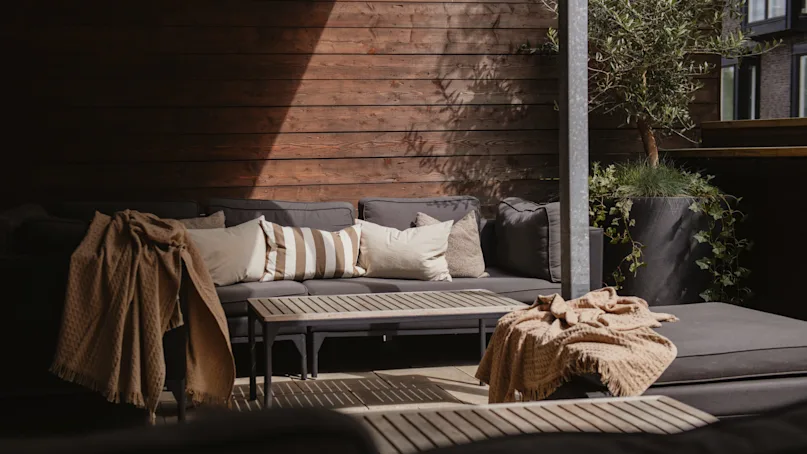 A cozy outdoor seating area featuring gray couches with beige cushions, wooden tables, and decorative blankets, set against a wooden fence and large potted plants. 