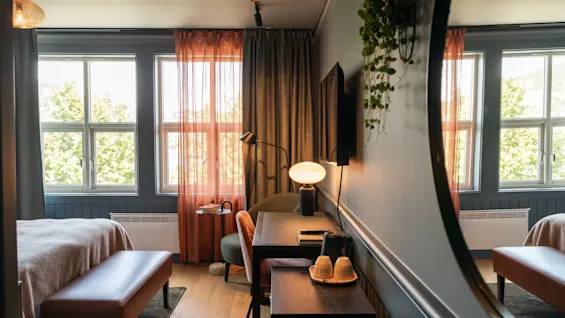 Et hyggeligt hotelværelse har en seng og et skrivebord; store vinduer med tynde gardiner lader naturligt lys strømme ind. Et spejl reflekterer en del af rummet og udsigten udenfor.
