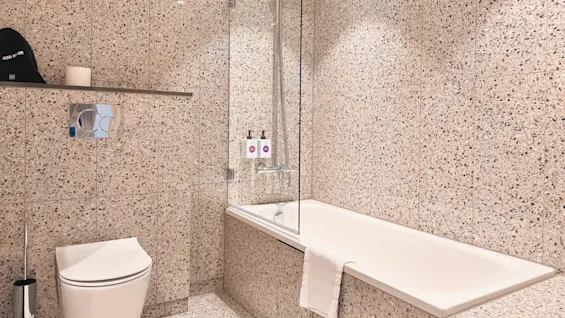 A bathroom with a wall-mounted toilet, a bathtub with a glass screen, and a showerhead. Speckled beige tiles cover the walls. A towel lies on the bathtub edge.