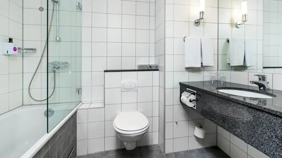 A bathroom with a bathtub and shower combo, toilet, and a granite sink counter adorned with towels and glasses. White tiles cover walls and floor, creating a clean, modern look.