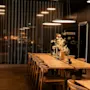 Et varmt og innbydende spise- eller møterom med et langt trebord, stoler og dekorative elementer, opplyst av moderne pendellamper.