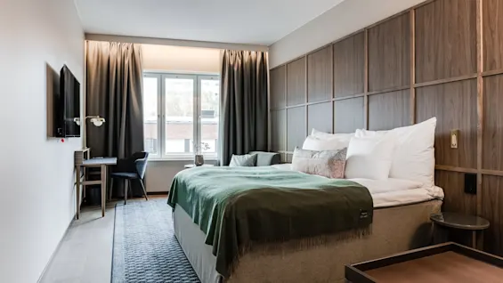 En dobbeltseng med grønne tæpper er centralt i hotelværelset. Et TV er monteret på venstre væg, og et vindue med gardiner giver naturligt lys.