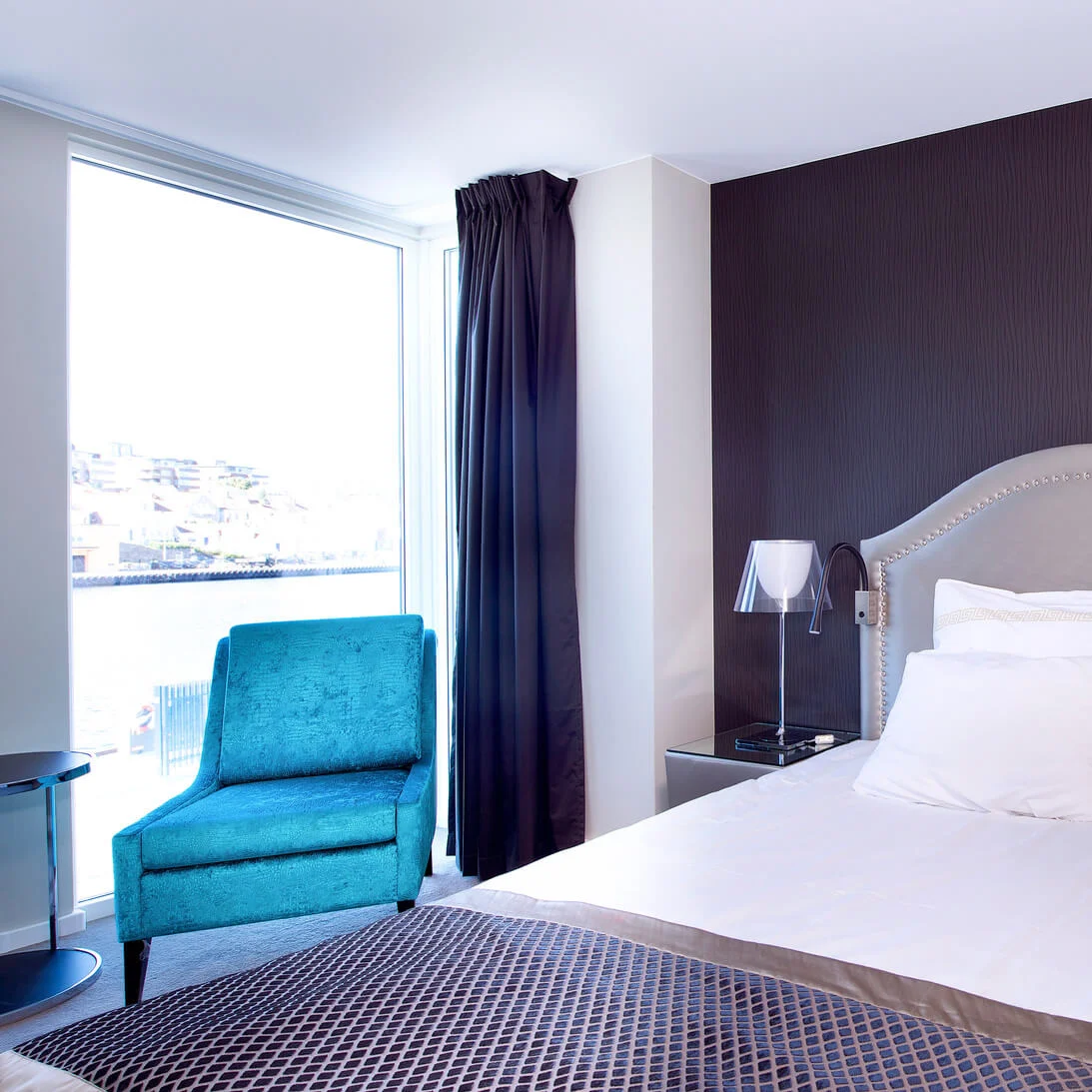 A neatly made bed with a blue armchair, under bright lighting, inside a modern bedroom with a large window.