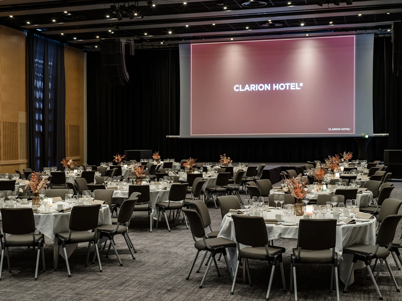 En rymlig evenemangshall med många runda bord och stolar, elegant dukade för en sammankomst. 