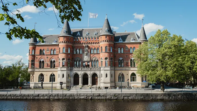 Storslått byggverk med røde mursteiner og buede vinduer, stående ved en elv under en klarblå himmel. Trær rammer inn scenen og gir naturlig grøntområde.