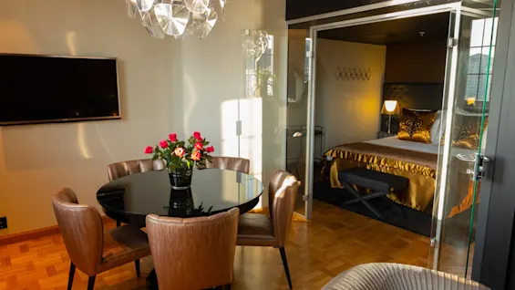 Et rundt spisebord med en vase med røde blomster er omgitt av skinnstoler, plassert i et moderne rom med en hengende lysarmatur og en seng synlig gjennom glassdører.