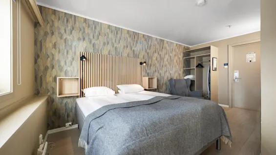 Lyst hotellrom med en oppredd dobbeltseng, mønstret fondvegg og tredetaljer.