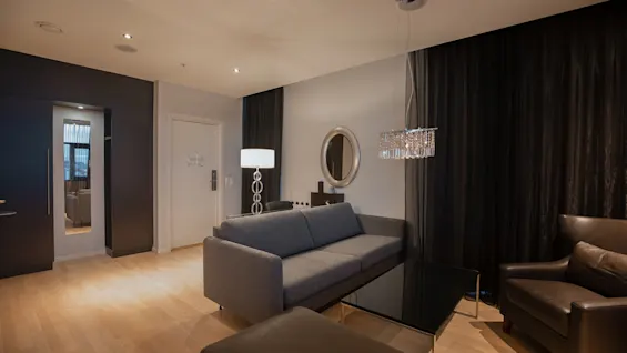 Grå sofa ved siden av et glassbord under en hengende lysekrone. Et rundt speil henger på veggen, med gardiner og en gulvlampe i nærheten.