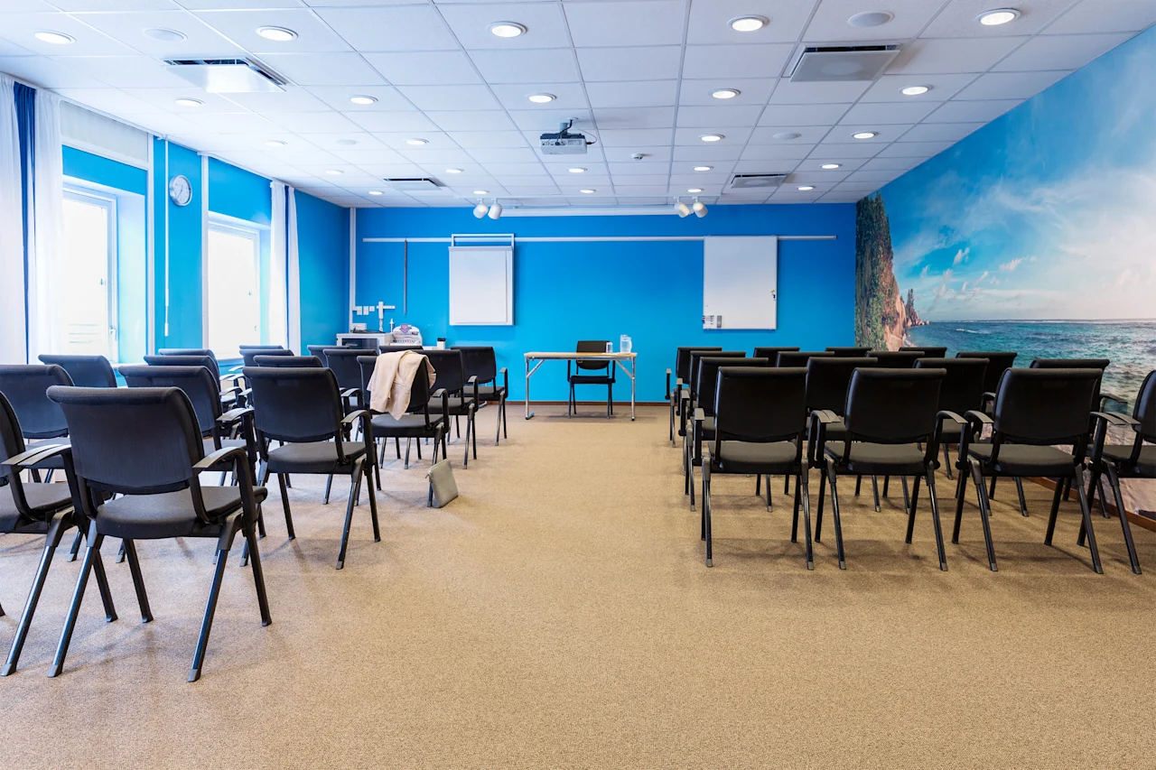 A spacious conference room with rows of empty black chairs facing a blue wall, featuring a flip chart and beach-themed mural, under bright ceiling lights. Windows line the left side.