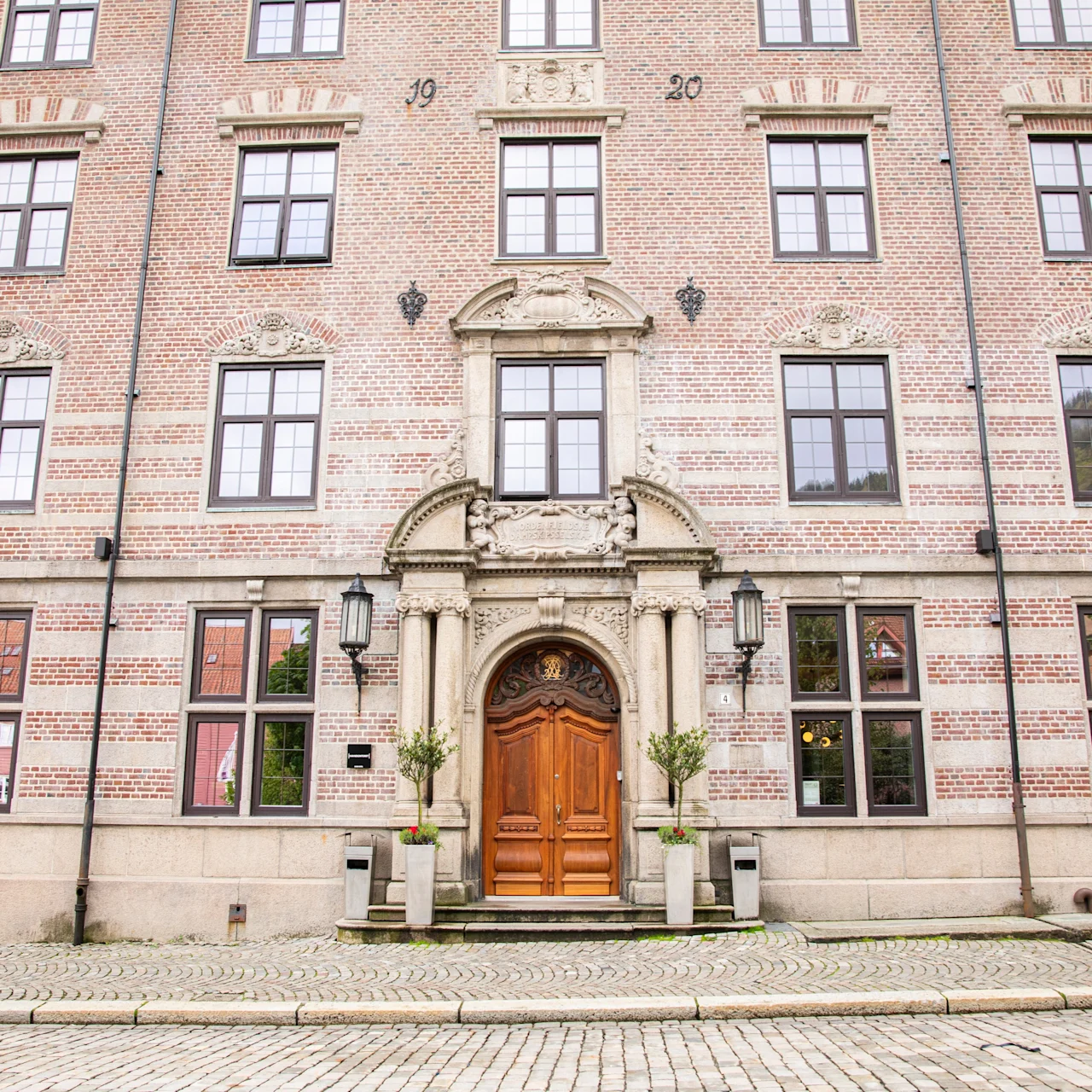 A brick building features a grand wooden door adorned with columns and plants, surrounded by large arched windows. The year "1920" is inscribed above, situated on a cobblestone street.