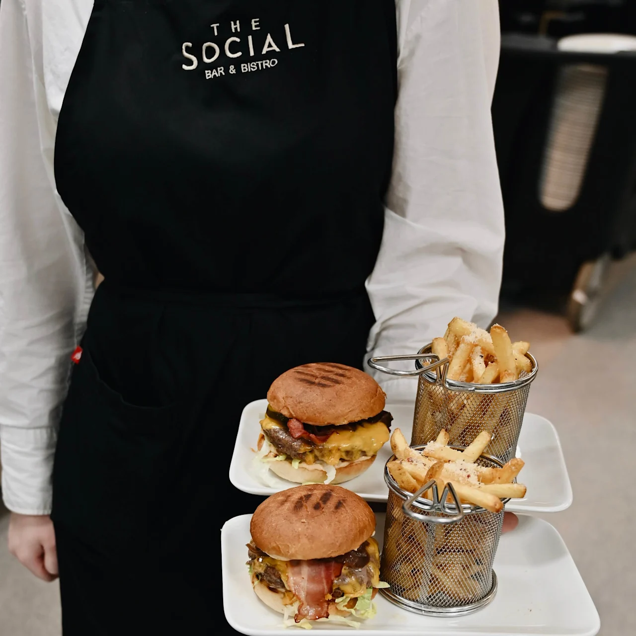 Servitör bär två tallrikar, varje med en hamburgare och pommes frites, medan de bär ett svart förkläde med texten "THE SOCIAL BAR & BISTRO." De står i en restaurangmiljö.