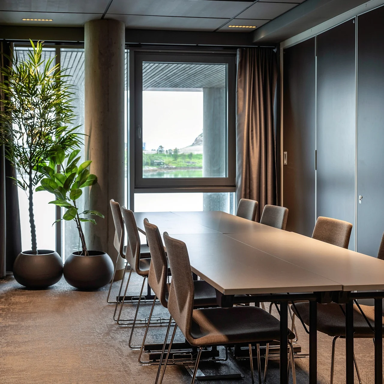 Et langt konferencebord er omgivet af stole i et moderne mødelokale med store potteplanter og et vindue med udsigt over udendørsmiljøet.