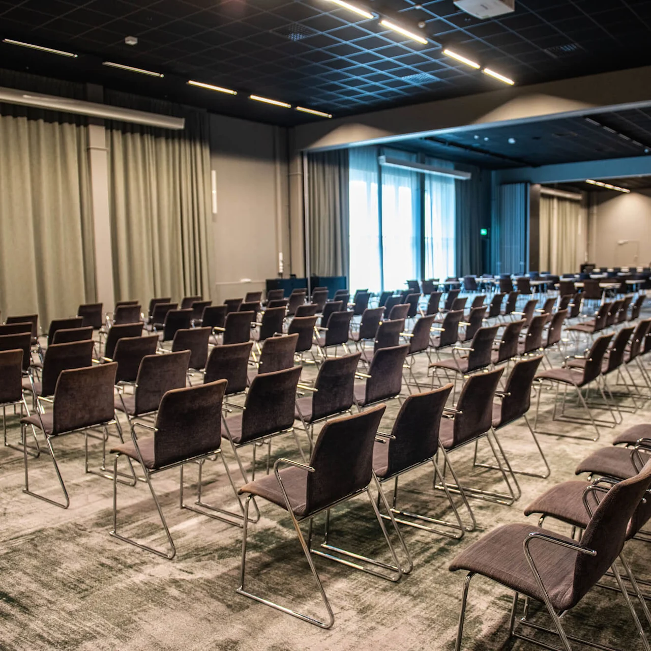 Rows of empty chairs face a stage in a large, carpeted conference room. The room features curtains, overhead lighting, and large windows providing natural light.