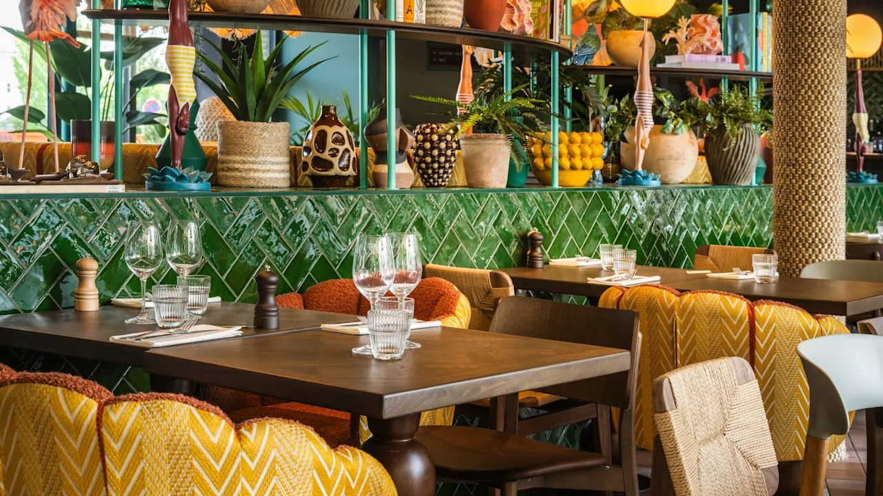 Borde med glas og servietter, omgivet af mønstrede stole i en farverig restaurant fyldt med planter og dekorative genstande på grønne fliser.